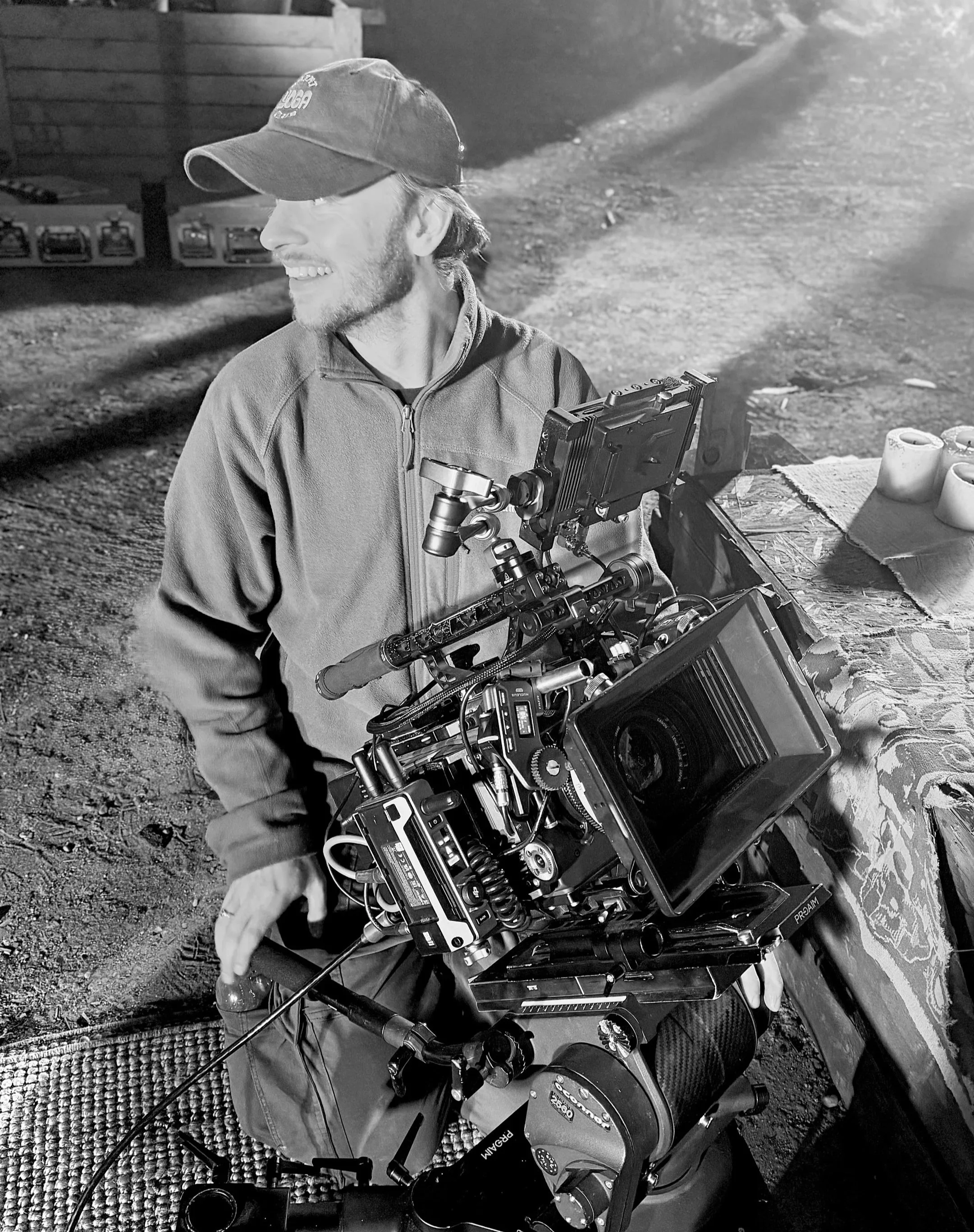 A man smiling, wearing a baseball cap and jacket, appears to be operating a professional camera mounted on a stabilizer rig outdoors at night.