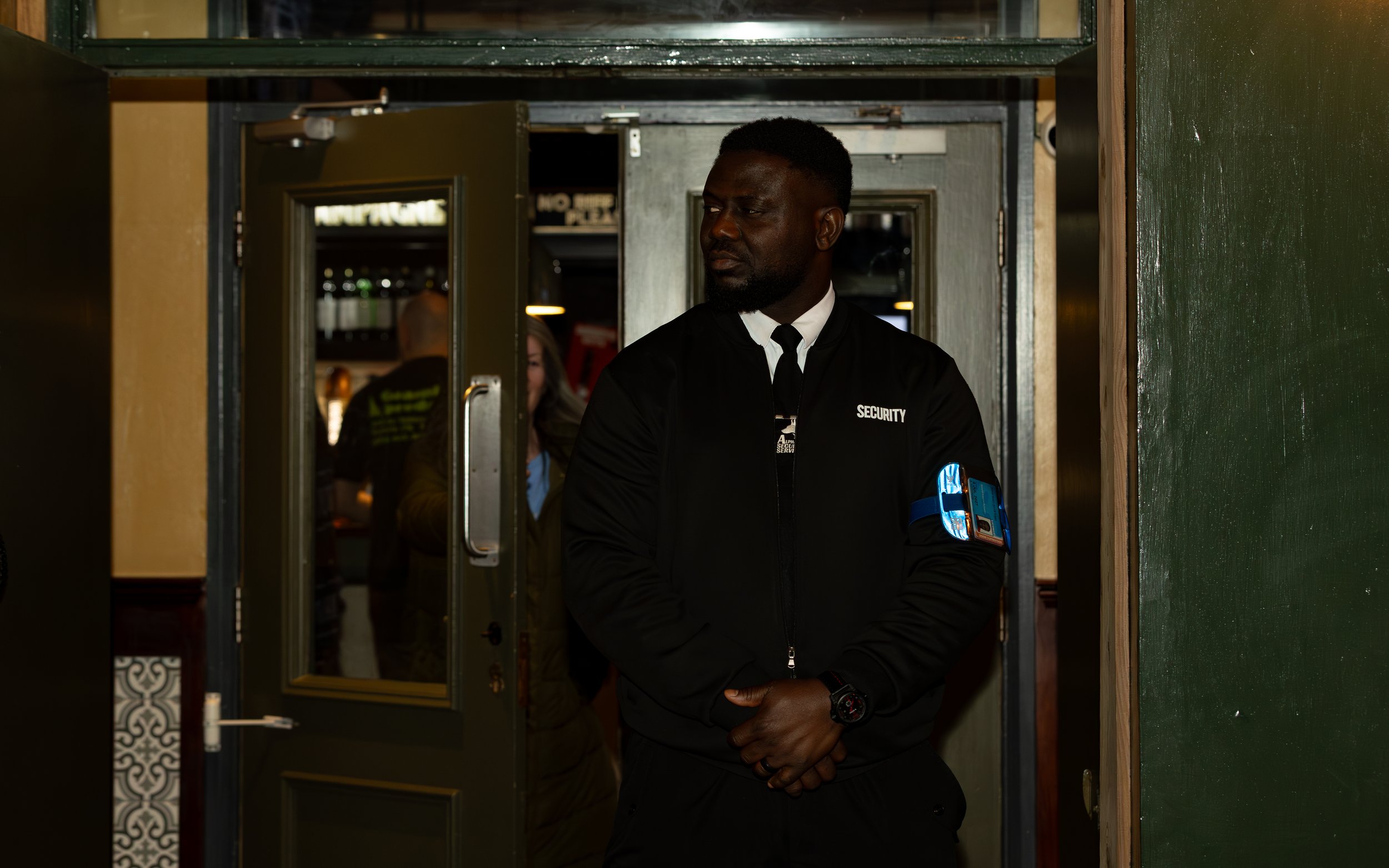 A security guard dressed in black uniform standing at a doorway, with a blue armband on his right arm.