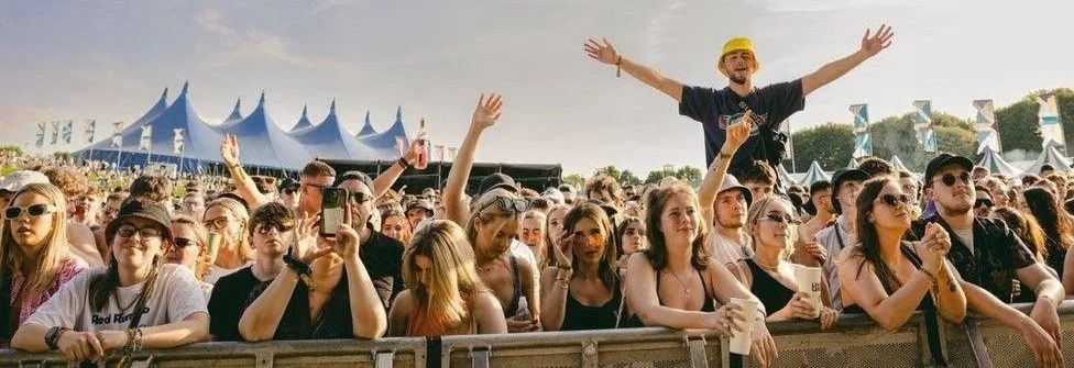 Crowd of young concert-goers at outdoor music festival with a man standing on a barrier with arms outstretched and wearing a yellow hat, tents in the background.