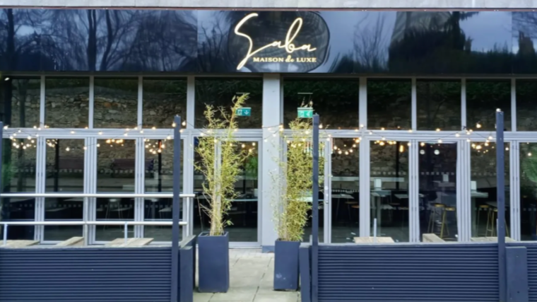 Storefront with a sign reading 'Ebera Maison de Luxe' above the entrance. The storefront has large glass windows, two potted plants outside, and string lights decorating the exterior.