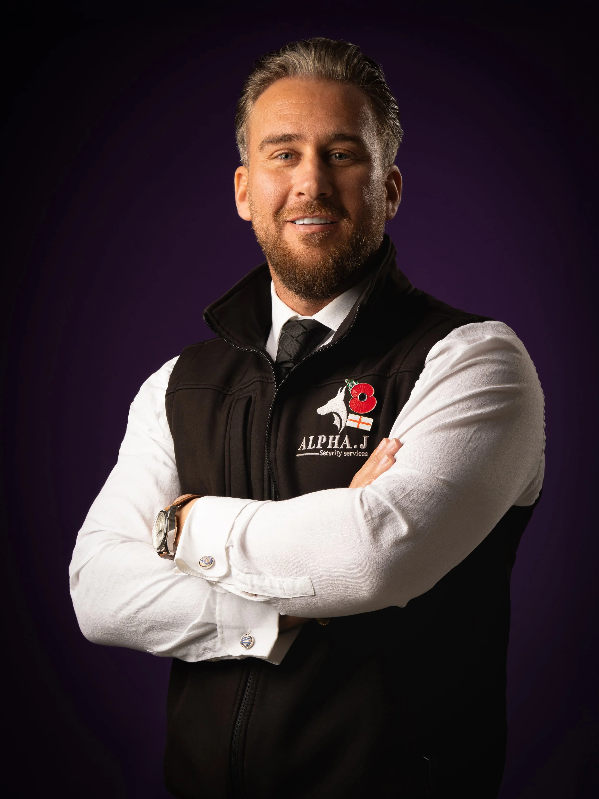 Portrait of a man with light skin, short gray hair, blue eyes, and a beard, wearing a white dress shirt, black vest with the logo of alpha J security services, a black tie, and a wristwatch, smiling with arms crossed against a dark purple background.