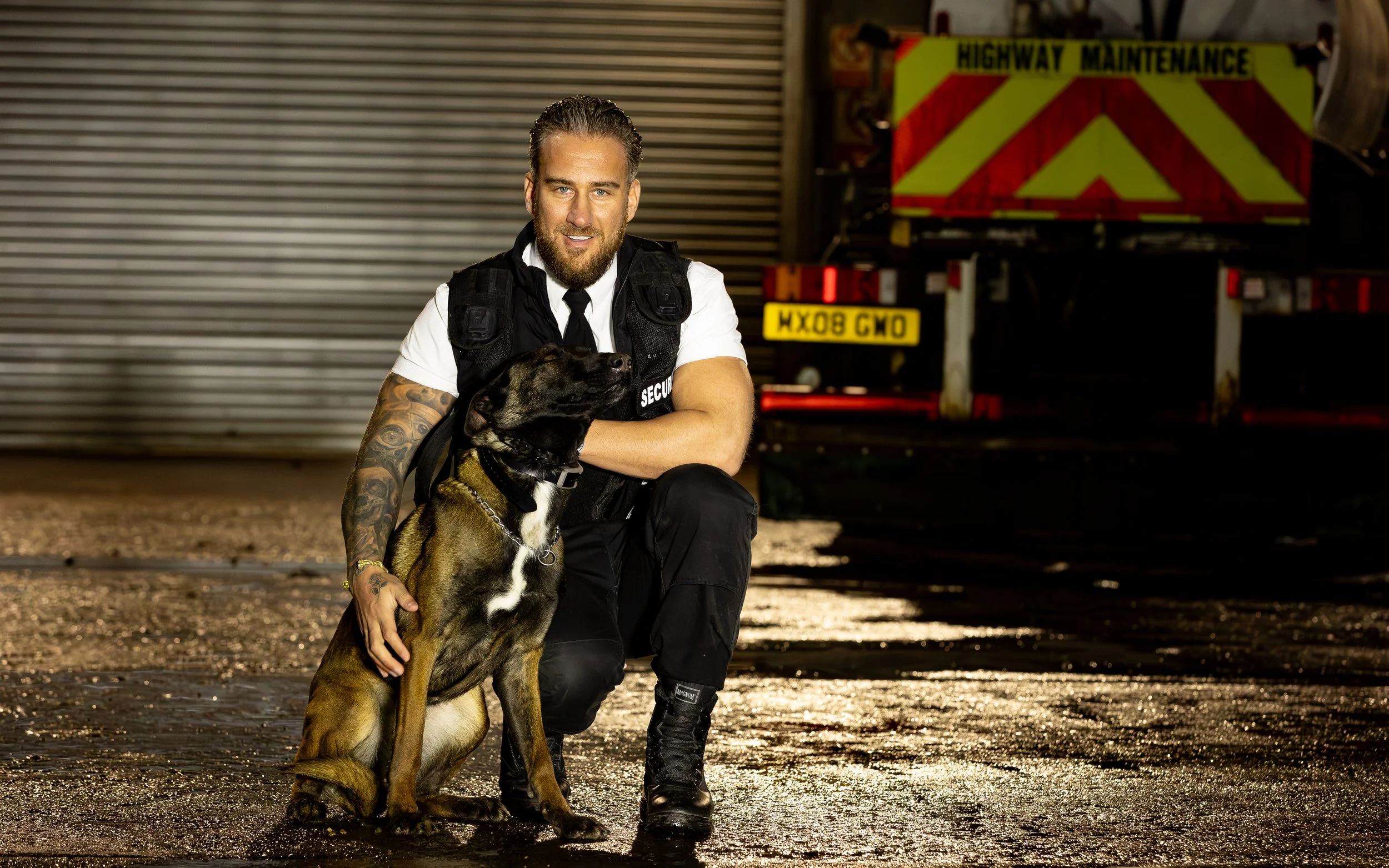 A man in security uniform crouching with a police dog in front of a closed metal shutter with a highway maintenance truck in the background.