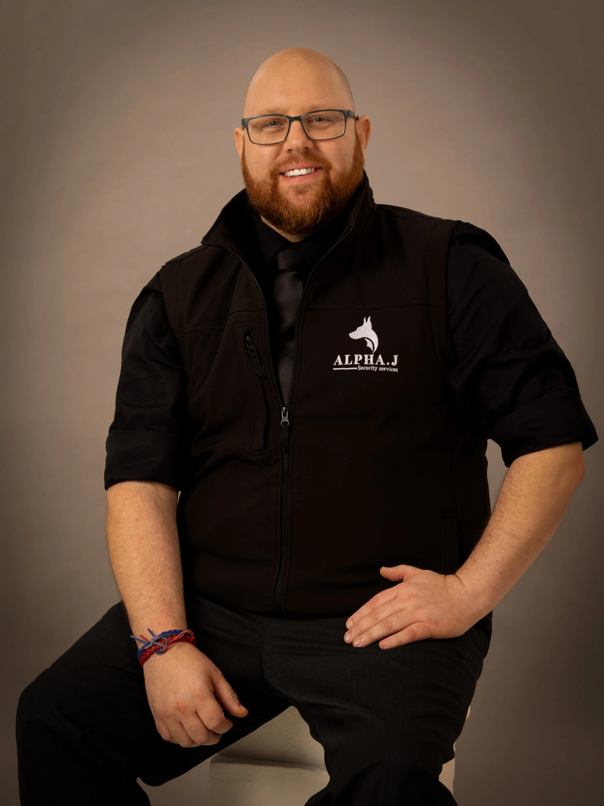 A man with a bald head, beard, glasses, wearing a black vest with a logo of a wolf head and the text "ALPHA.J Security services," black shirt, and black tie, sitting against a plain background.