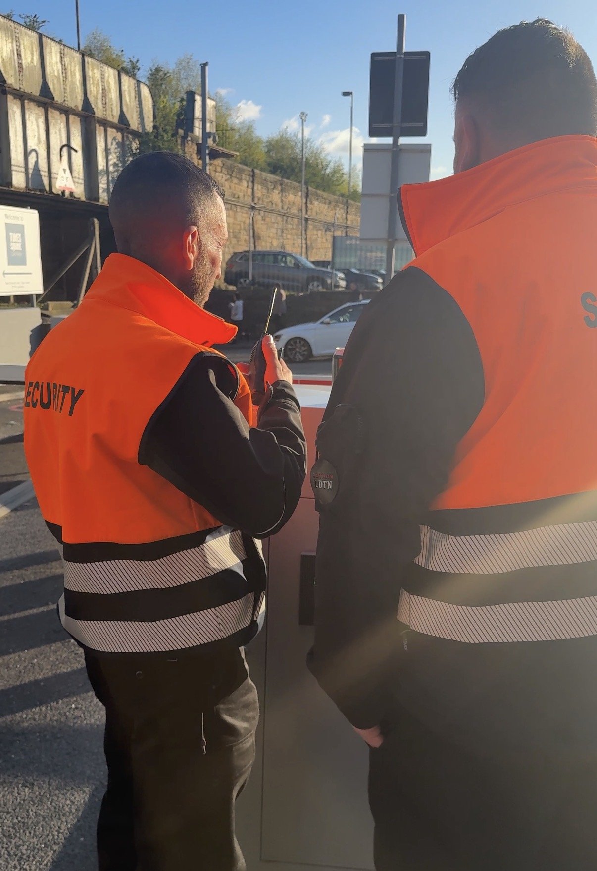 Two security personnel in orange vests standing outdoors near a parking lot, one of them is holding a walkie-talkie. The background includes cars, a brick wall, and a blue sky.