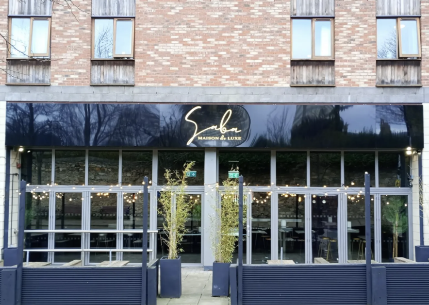 Exterior of a modern restaurant or cafe with a black awning displaying the name 'Sabor Maison de Luxe' in elegant gold lettering. The outdoor seating area features string lights and potted plants, with large windows reflecting trees and sky.