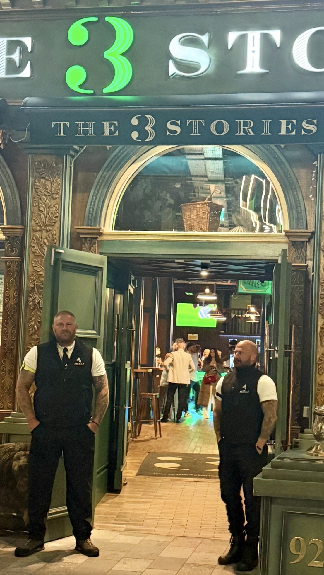 Interior of a bar or pub with people socializing and watching TV screens, two security guards standing at the entrance, ornate wooden decor, and a sign that reads 'The 3 Stories'.