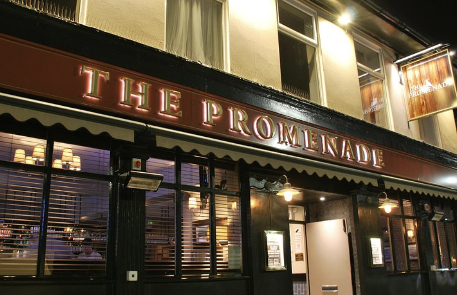 Nighttime exterior view of The Promenade restaurant with illuminated red sign and interior visible through windows.