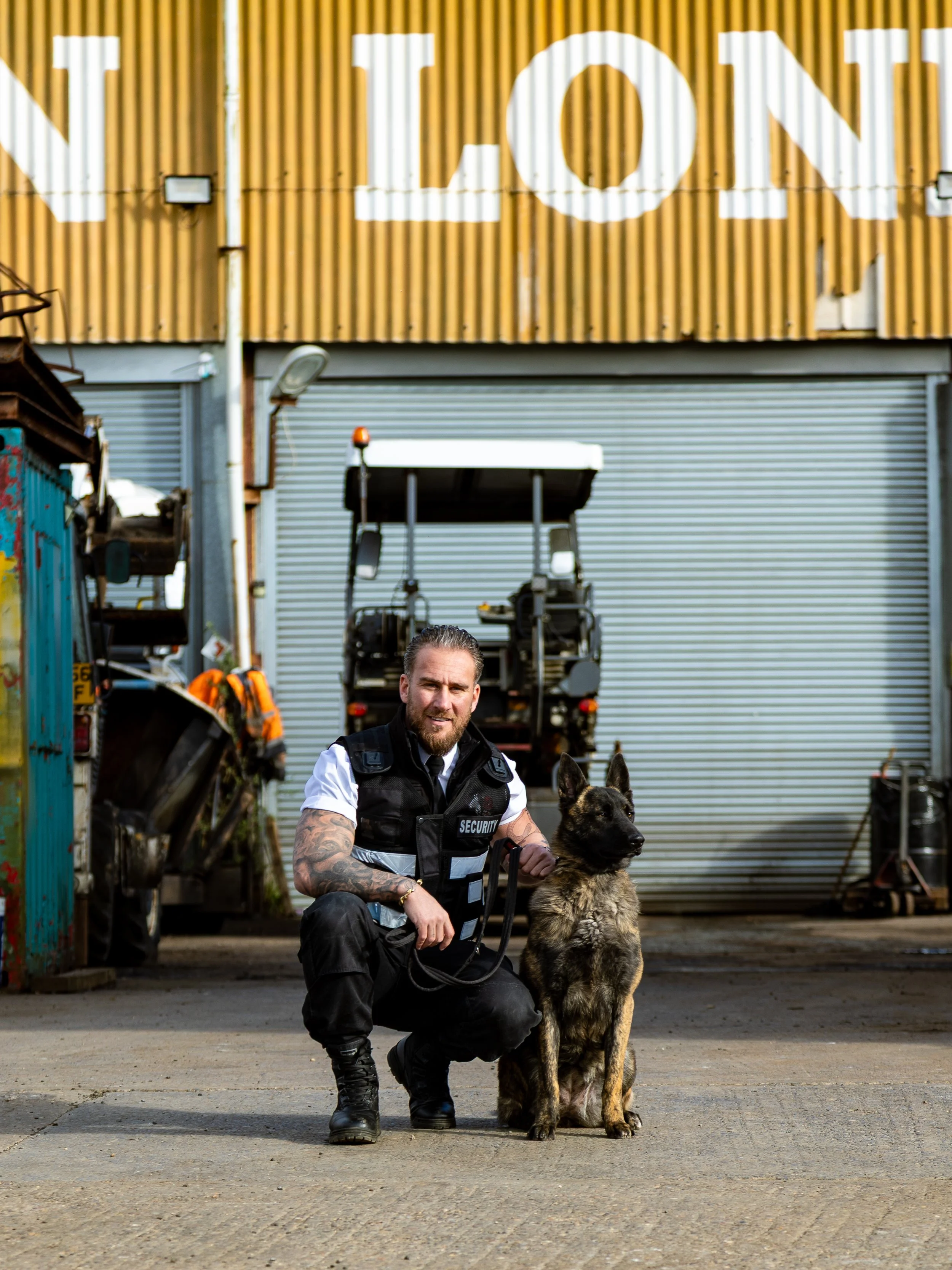 Kneeling security guard in black uniform with tattoos and a German shepherd dog outside a large industrial building with yellow corrugated metal walls.