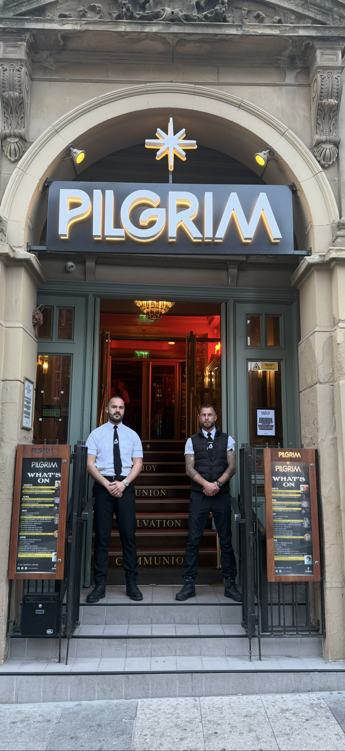 The entrance of a building named PILGRIM with a neon sign. Two men are standing at the entrance, one with a white shirt and black tie, the other with a black vest and tattoos on arms. There are menus on stands on each side of the stairs.