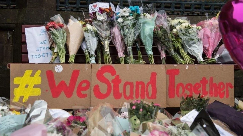A memorial with bouquets of flowers placed on a bench and a cardboard sign that reads '#We Stand Together' in red and yellow letters.