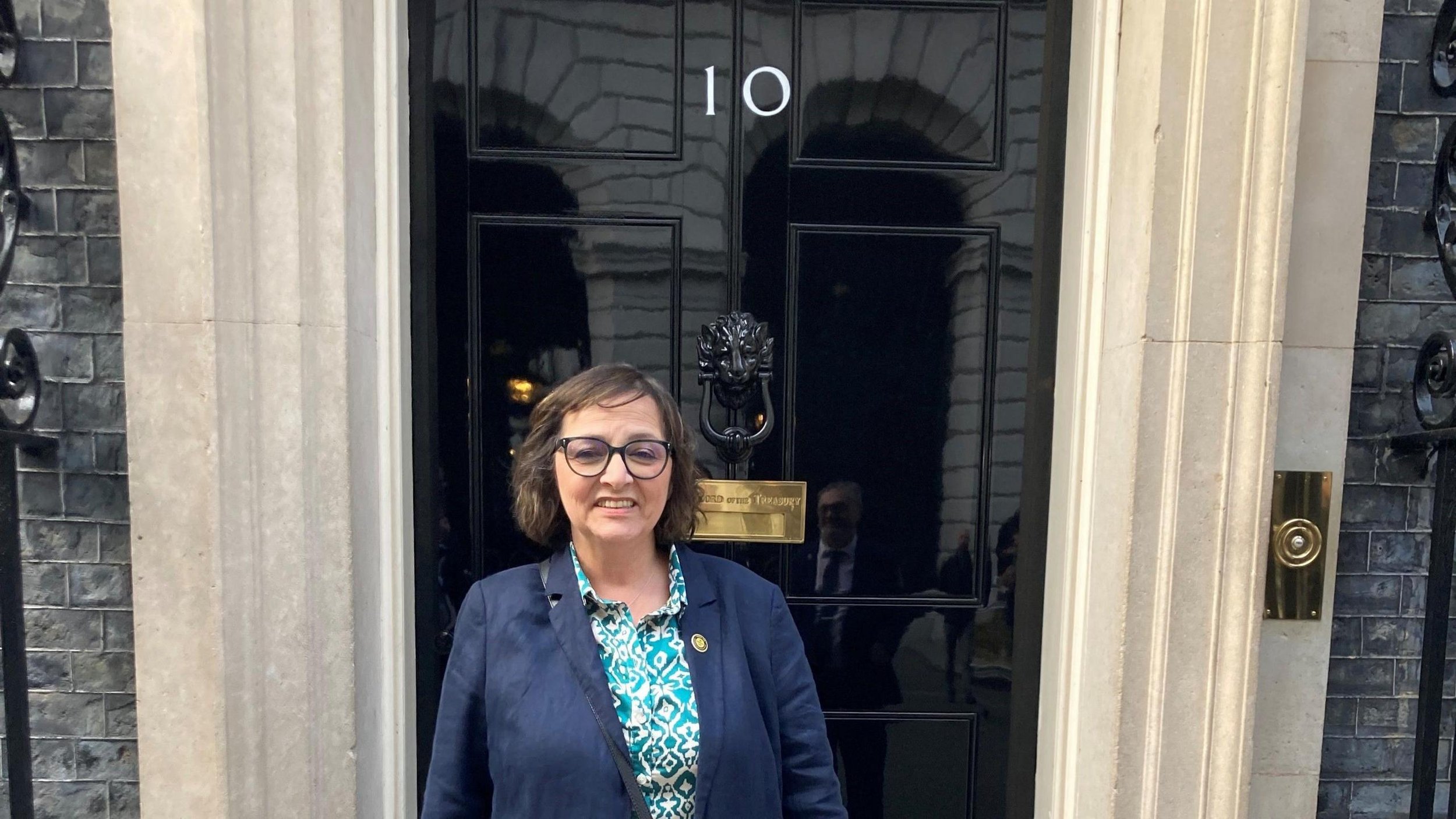 Woman with glasses and a patterned shirt standing in front of a black door with a lion head door knocker, the door has the number 10 and a brass letterbox.
