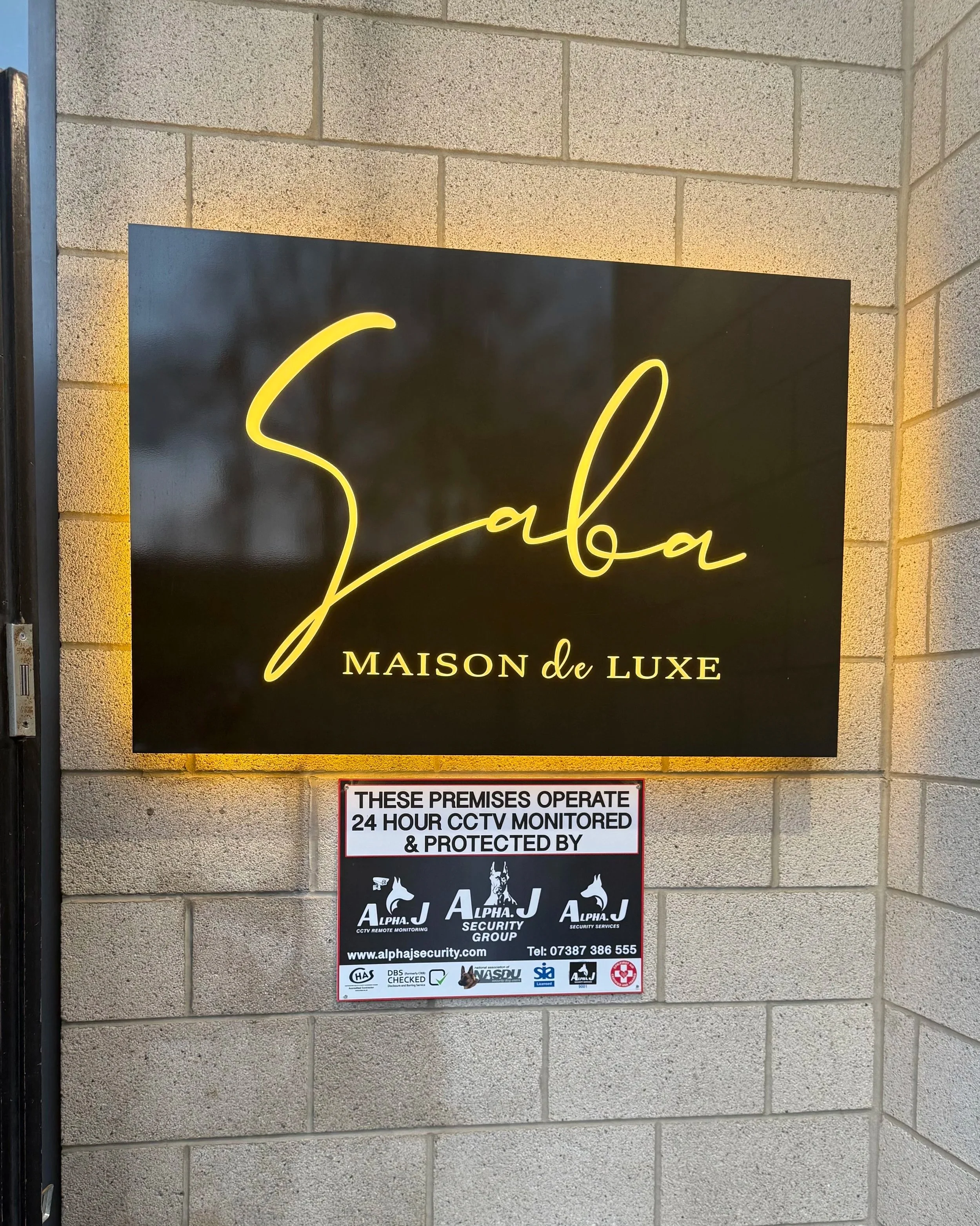Photo of two signs on a wall. The top sign is black with yellow cursive text saying 'Salba' and smaller text underneath reading 'MAISON de LUXE'. The bottom sign is rectangular with a white background, black text that states 'THESE PREMISES OPERATE 24 HOUR CCTV MONITORED & PROTECTED BY' and logos for security services.
