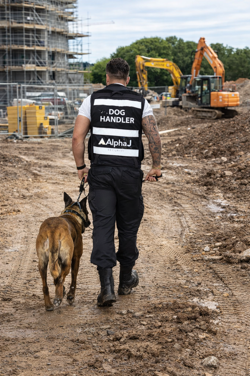 A security dog handler walking with a brown dog at a construction site with heavy machinery and building scaffolding.