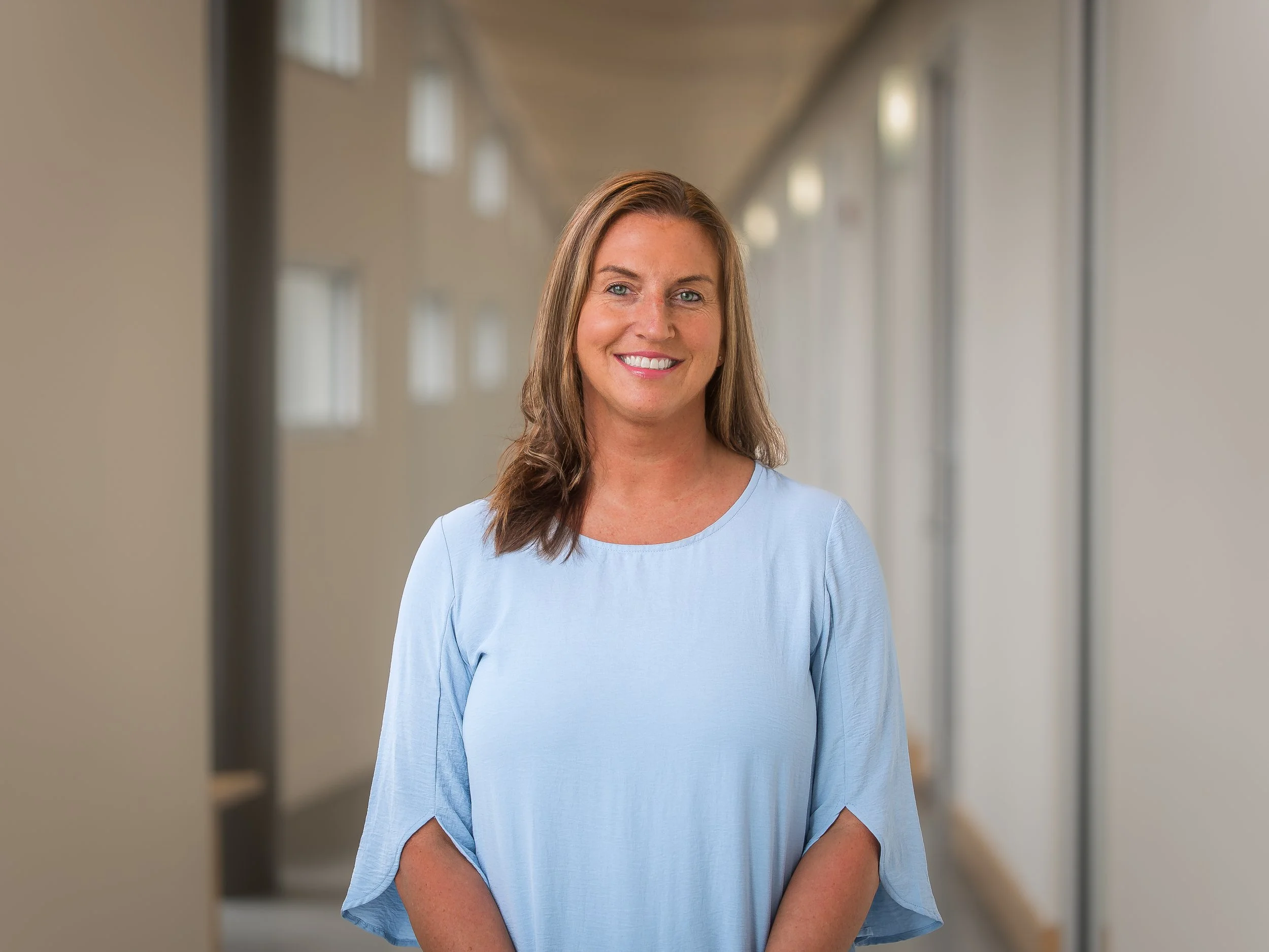 A smiling woman with shoulder-length brown hair, wearing a light blue blouse, standing in a bright corridor with windows along the side.