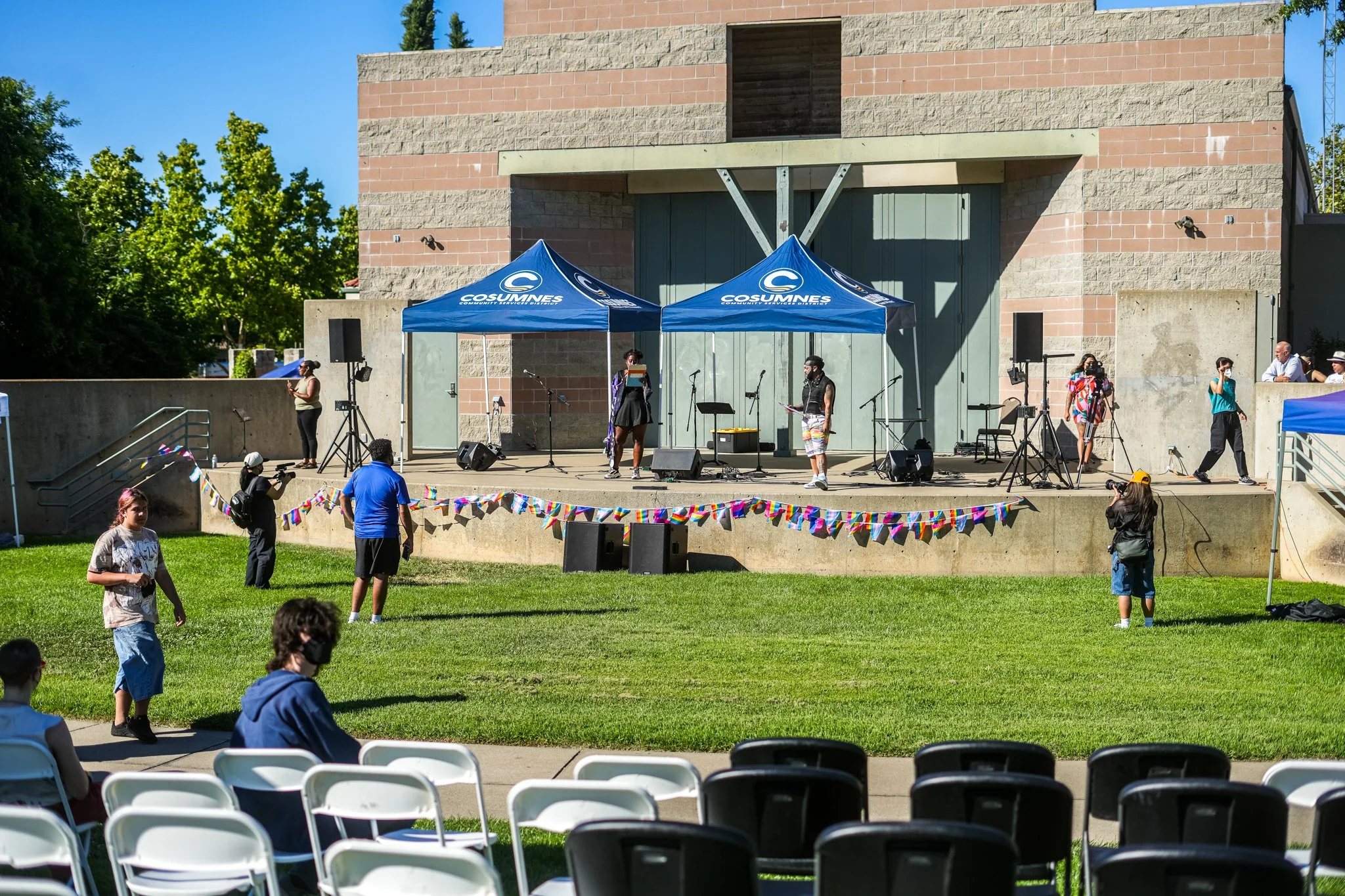 Outdoor community concert with professional live sound system, stage speakers, microphones, pop-up tents, and audience seating in a park setting.