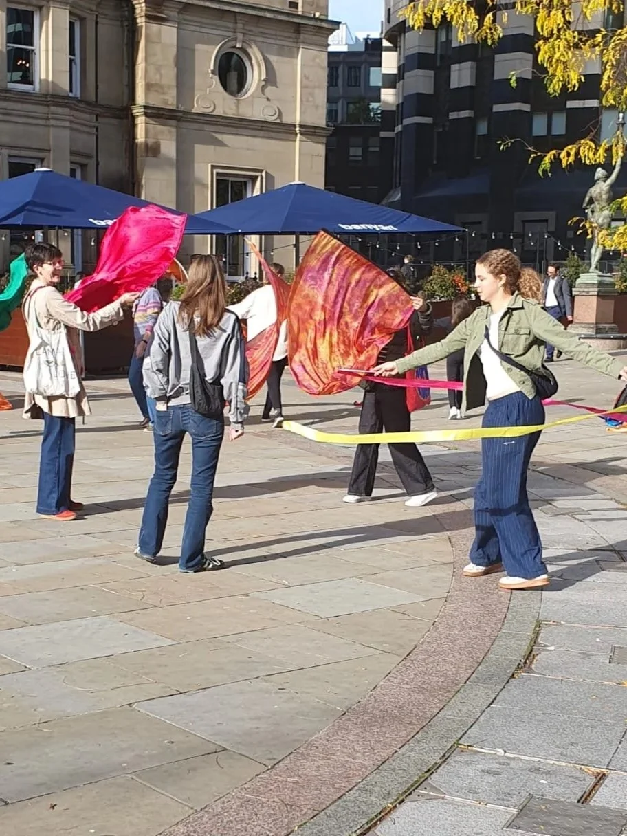 A group of women are waving flags and ribbons and dancing in the city centre on a sunny day