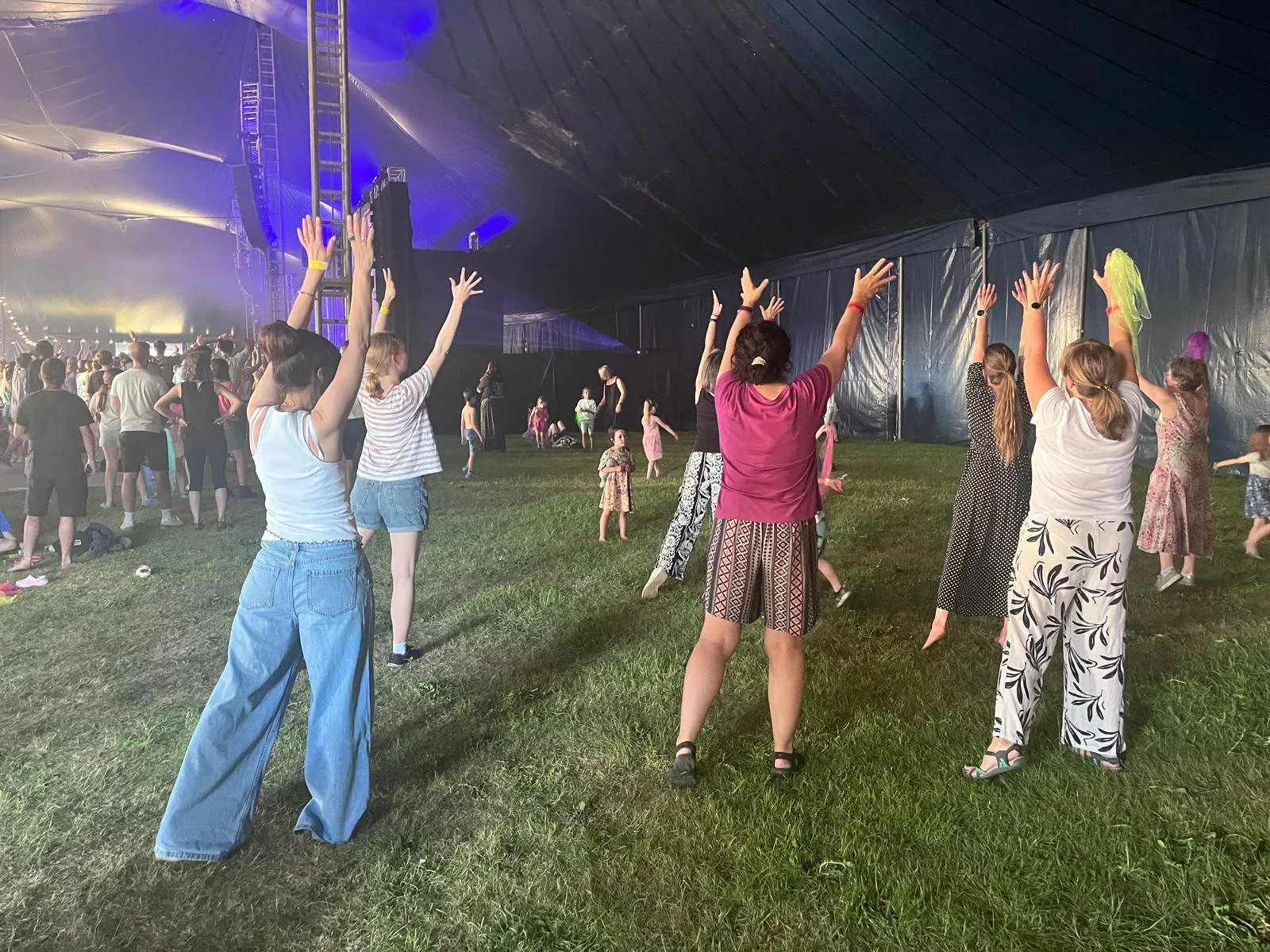 A group of women in a large tent with grass on the ground stand facing towards a stage with their arms raised.