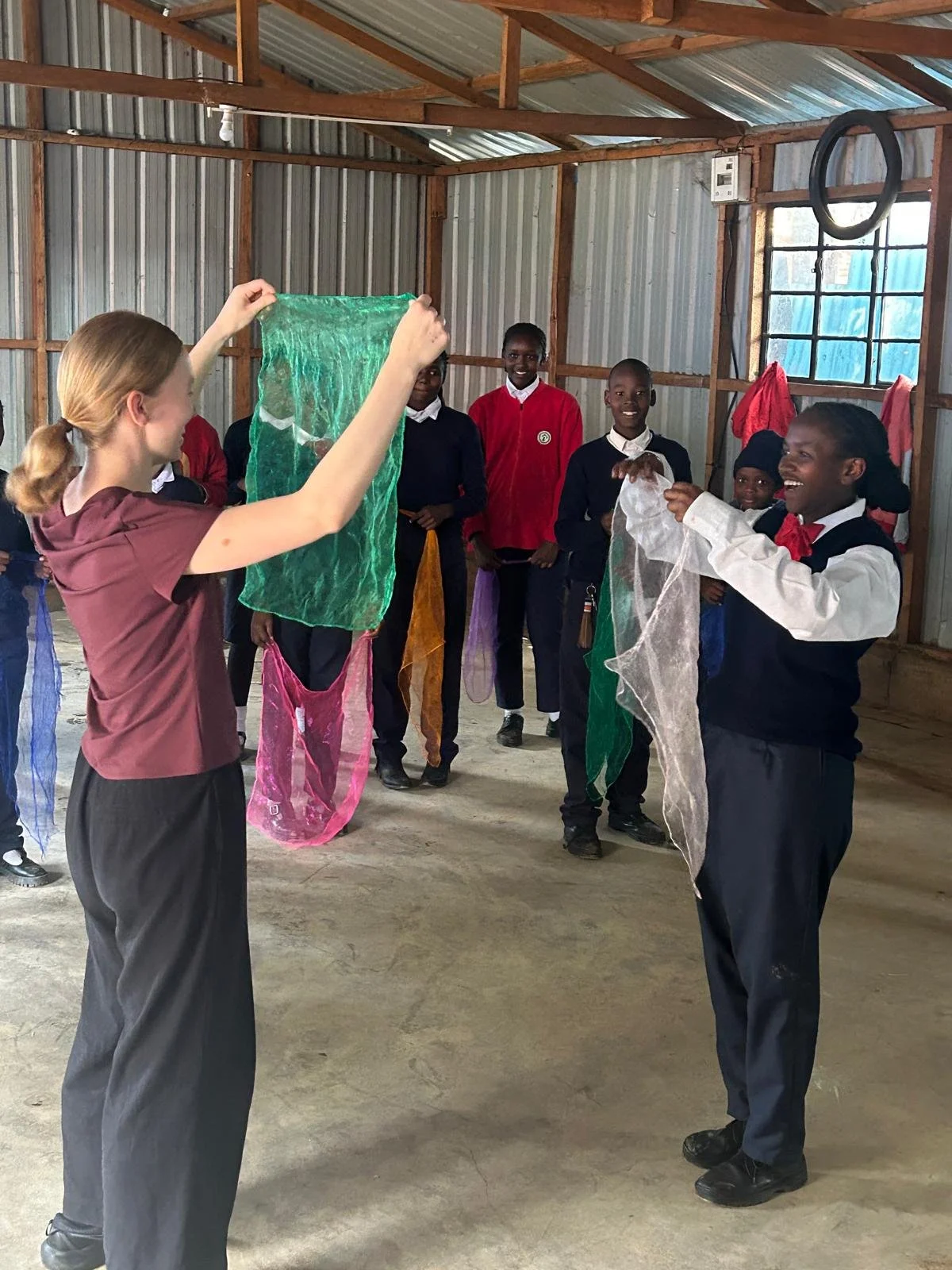 Two people face each other lifting up scarves to mirror each other. A group of young people stand by and watch, smiling. 