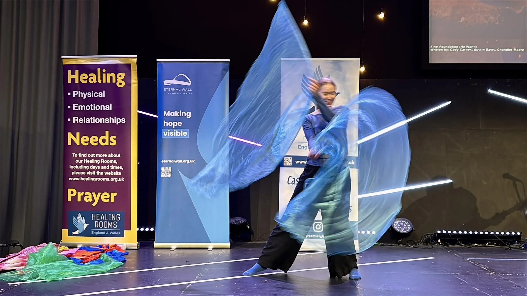 A woman with two blue, sheer flags, dances on a stage in worship.