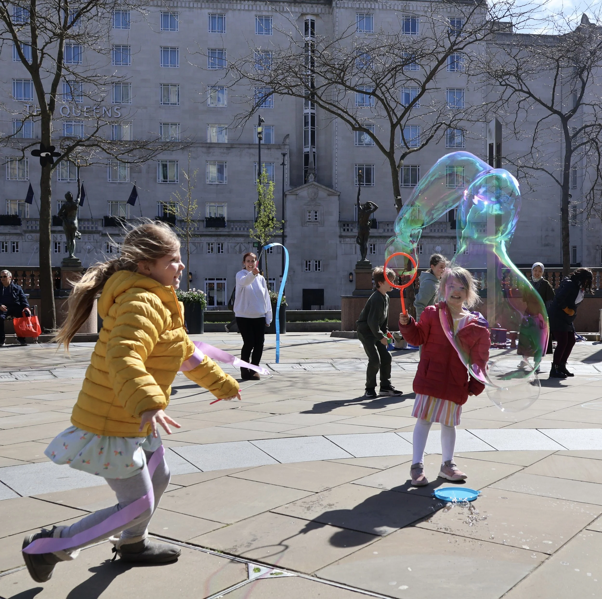 A child with a pink coat is making a large bubble with a giant bubble maker. Another child in a yellow coat runs and play on the streets in a city centre. It is very sunny