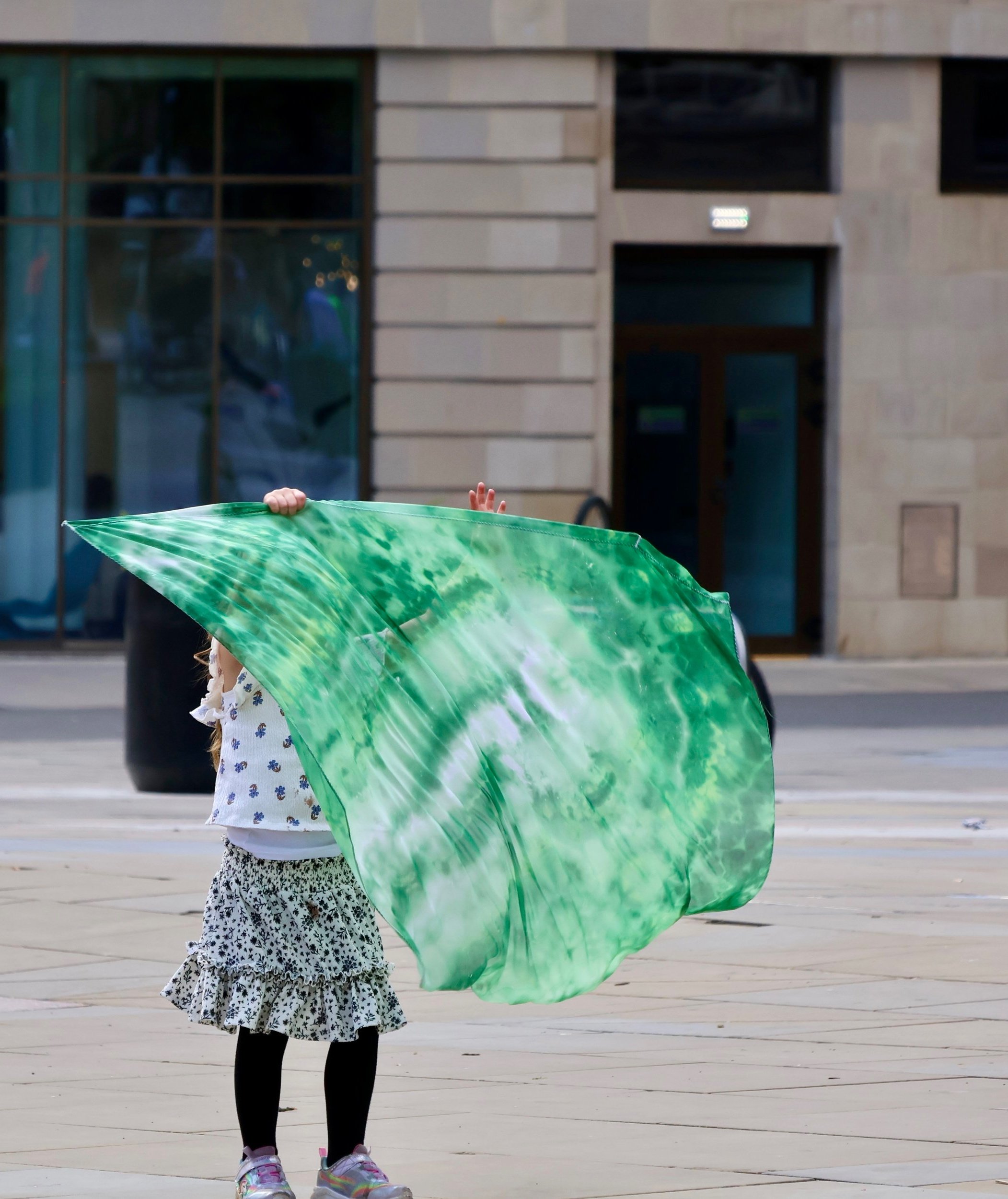 A small child standing on the street holding a big green flag up that covers their face.