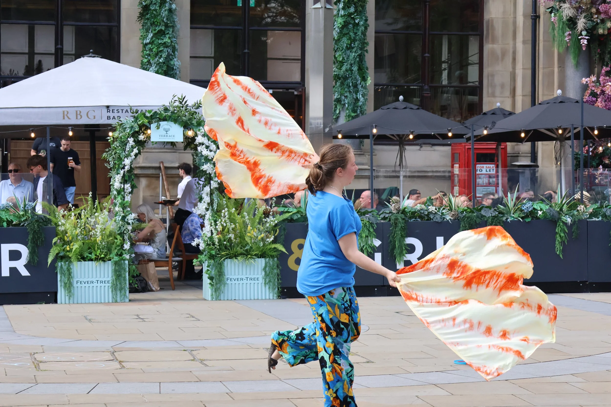 A woman with two yellow and orange flags dances on the streets in a city centre.