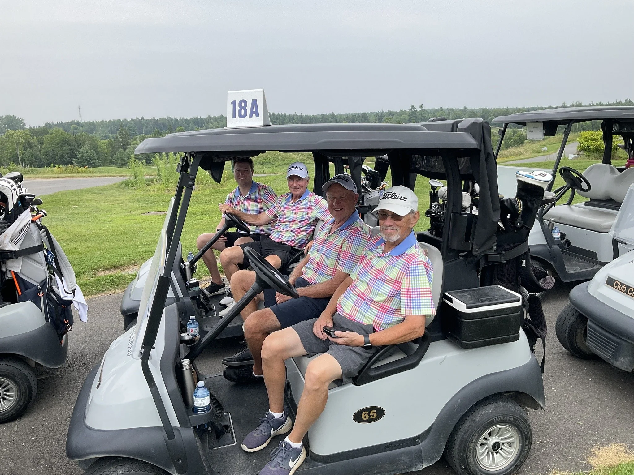 Four men in matching colorful shirts sitting in a golf cart with a sign "18A", surrounded by golf gear.