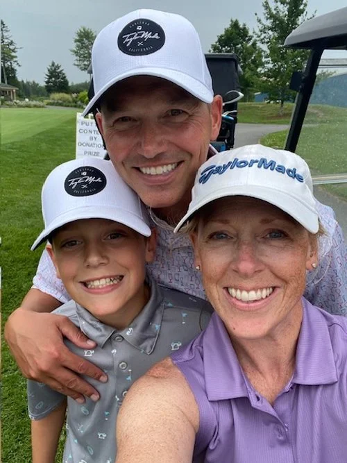 A smiling group of three people, two adults and a child, taking a selfie on a golf course. All are wearing caps, and the background shows greenery and a golf cart.