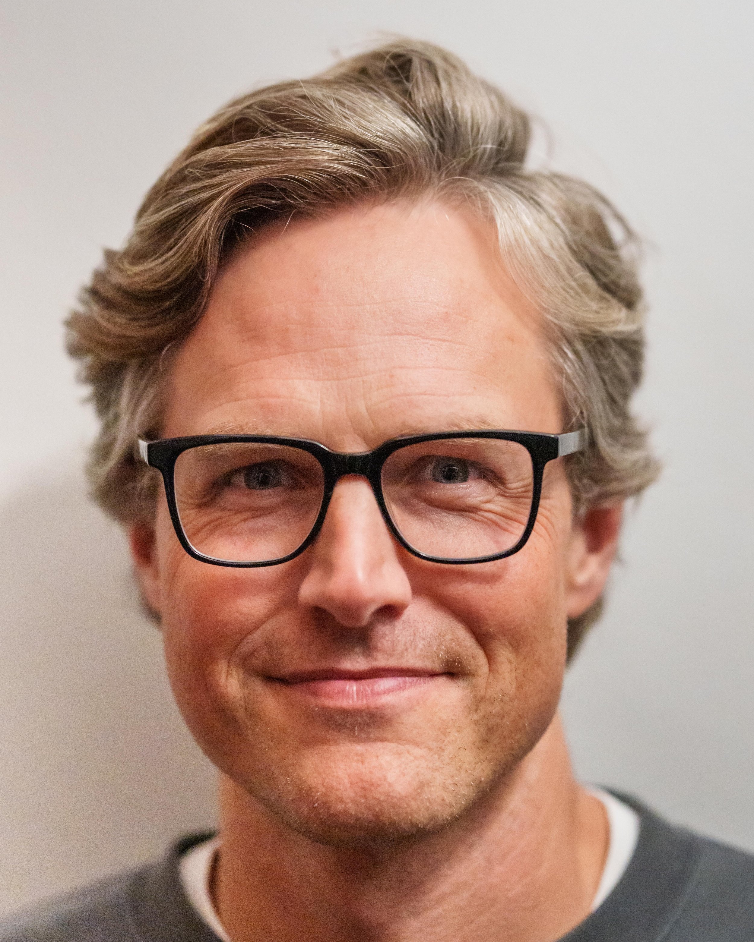 A middle-aged man with wavy gray hair, wearing black rectangular glasses, smiling slightly, against a neutral background.