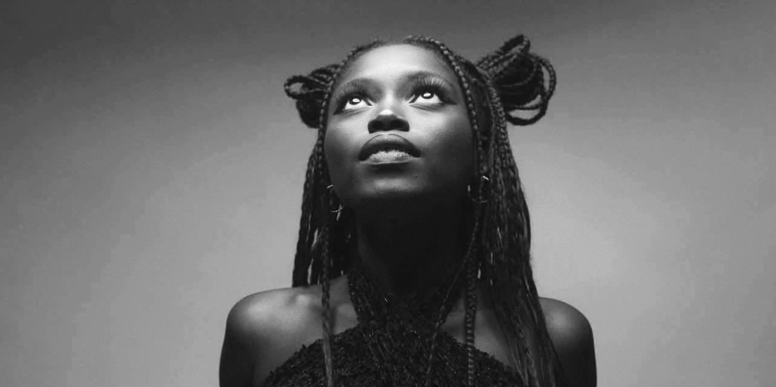 A black-and-white photo of a woman with braided hair looking upwards, with a neutral background.