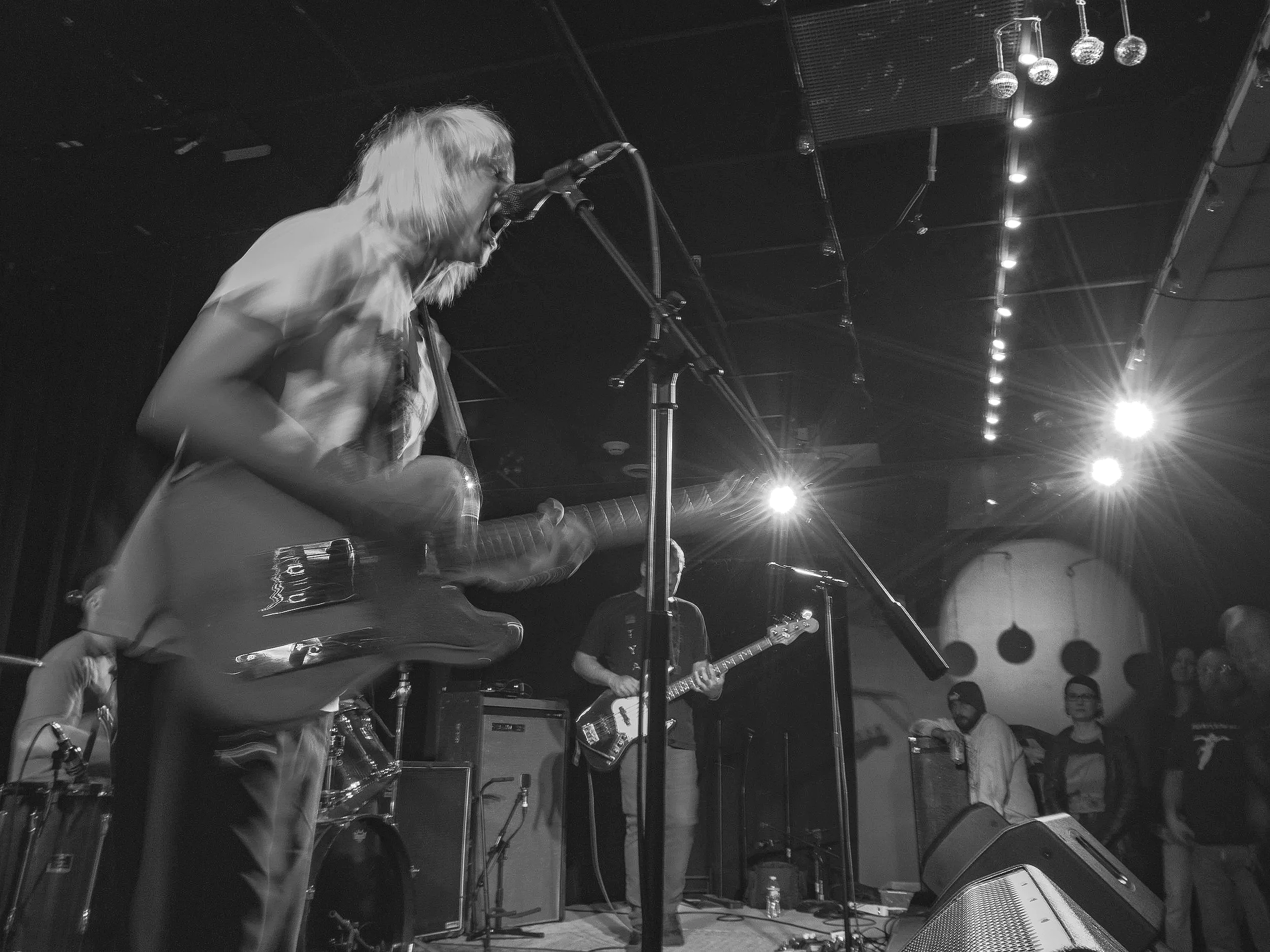 A black and white photo of a live music performance featuring a male musician with long hair playing an electric guitar and singing into a microphone on stage with his eyes closed. Multiple musicians are visible in the background along with music equipment, microphones, and stage lighting.