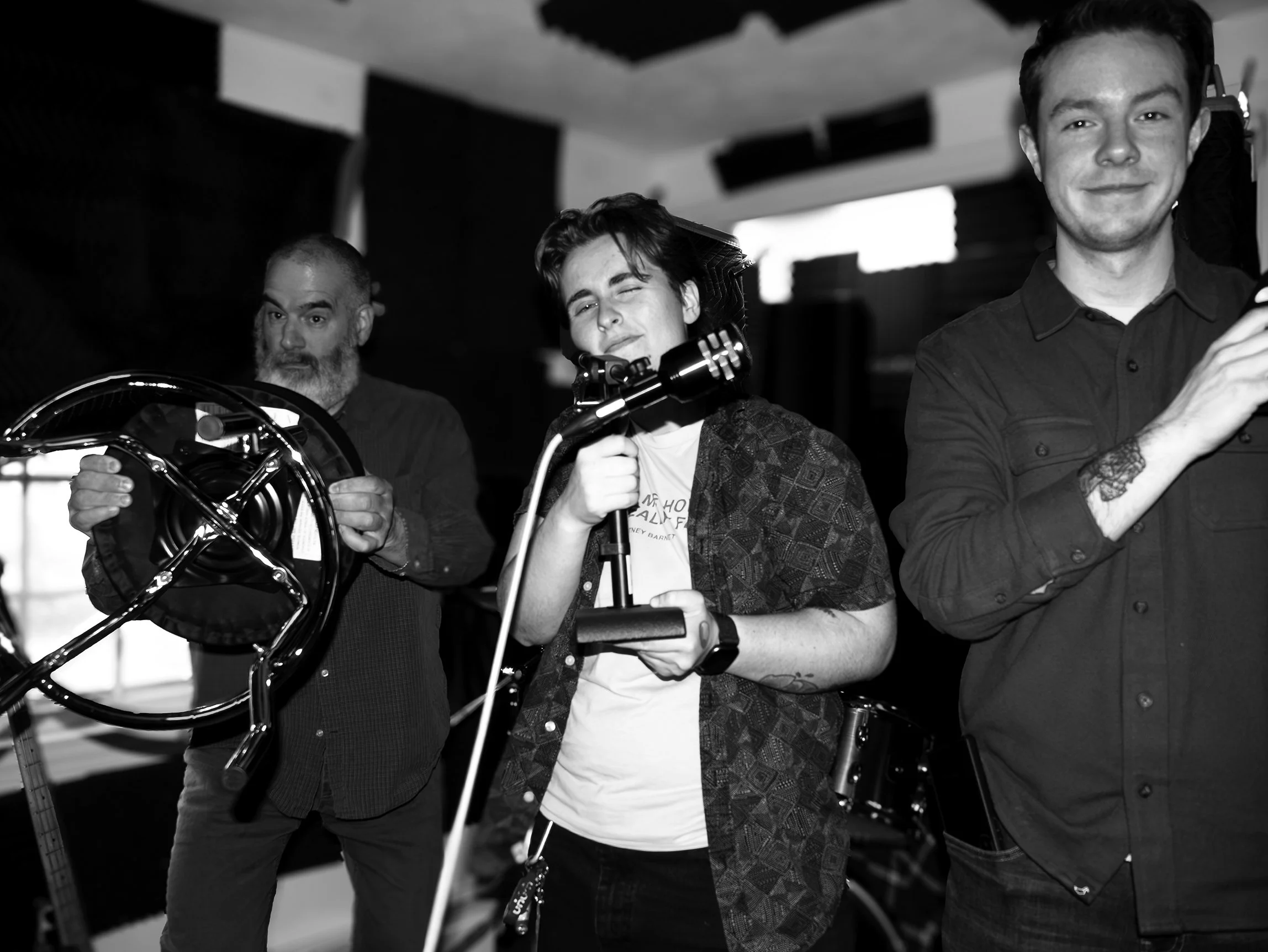 Three men in a music studio with black soundproofing panels, one holding a motorcycle helmet, one adjusting a microphone, and another smiling with arms crossed.