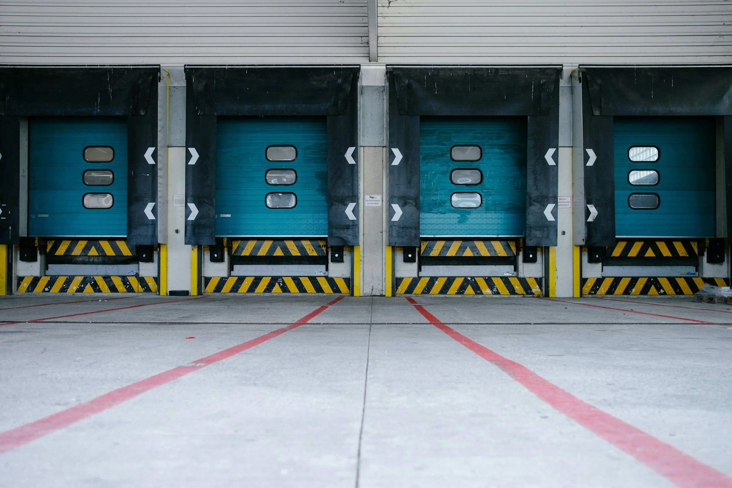 Multiple closed blue loading dock doors in a warehouse with black and yellow safety barriers and red parking lines on the concrete floor.