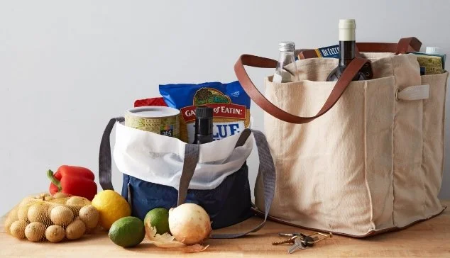 Two reusable shopping bags filled with groceries, including bottles, chips, and other food items, with fresh produce like potatoes, onions, and limes placed in front on a wooden surface.
