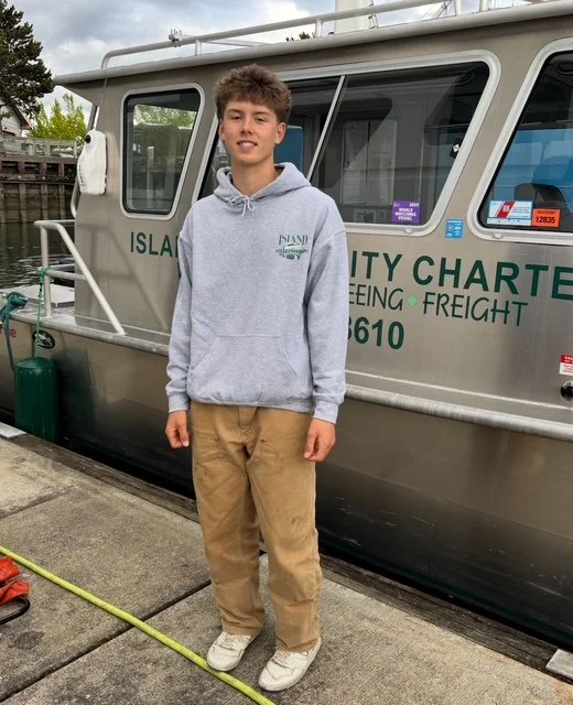 Young man standing on a dock in front of a boat, wearing a gray hoodie and khaki pants.