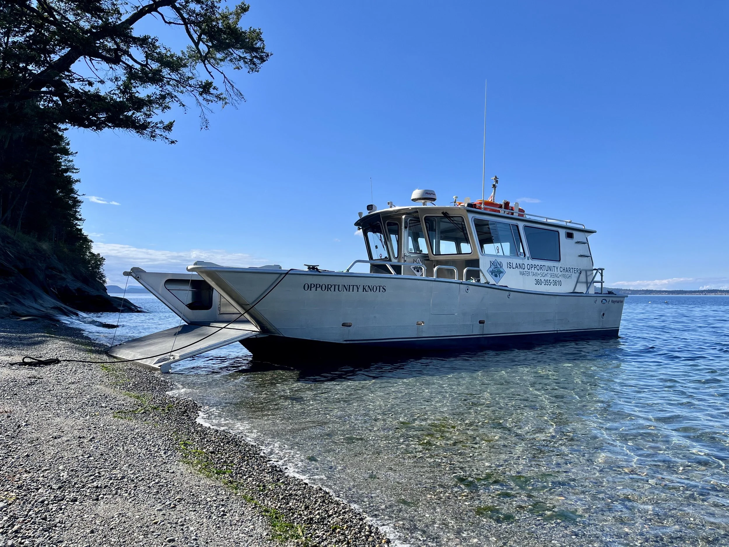 A boat named Opportunity Knots anchored near a rocky shoreline under a clear blue sky.