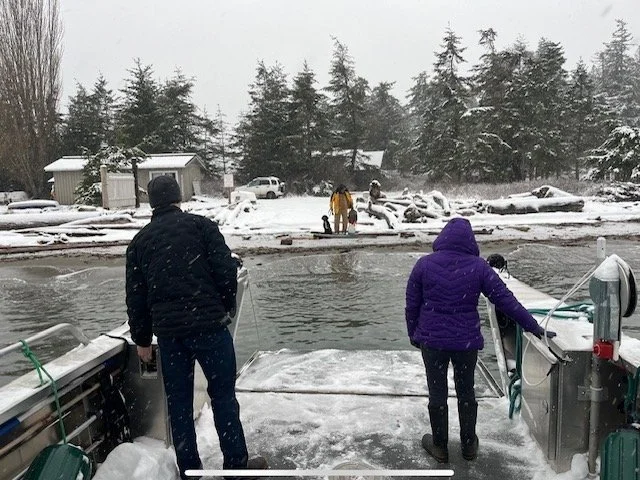 People in winter clothing launching a boat into a snowy lake with snow-covered trees and vehicles in the background.