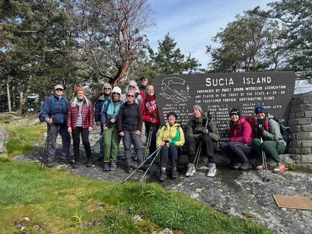 Group of hikers posing beside a sign that says 'Sucia Island' on a trail in a forested area.