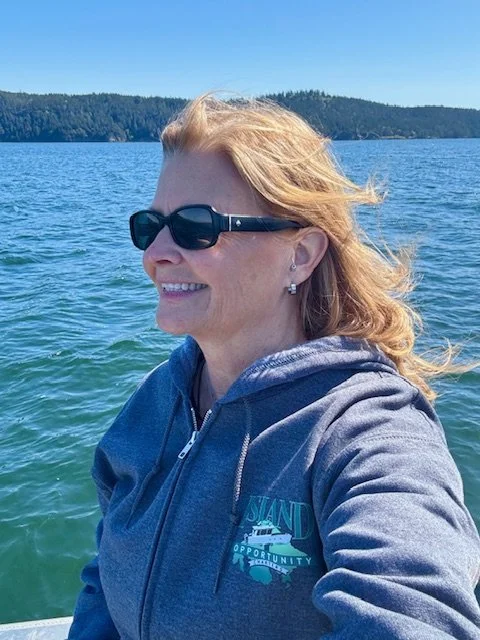 A woman with red hair wearing sunglasses and a blue hoodie, smiling, on a boat in a large body of water with a forested landmass in the background.