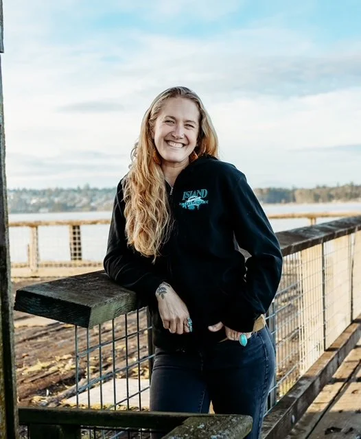 A smiling young woman with long wavy blonde hair leaning on a wooden railing outdoors with water and a distant shoreline in the background, wearing a black hoodie and jeans.
