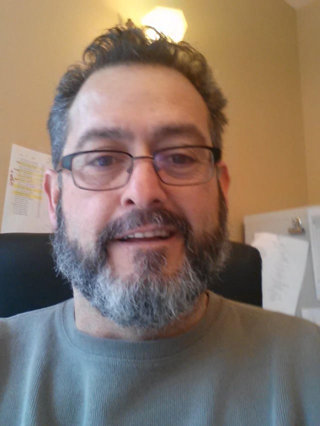 A middle-aged man with glasses, a beard, and short curly hair smiling at the camera in an indoor setting.