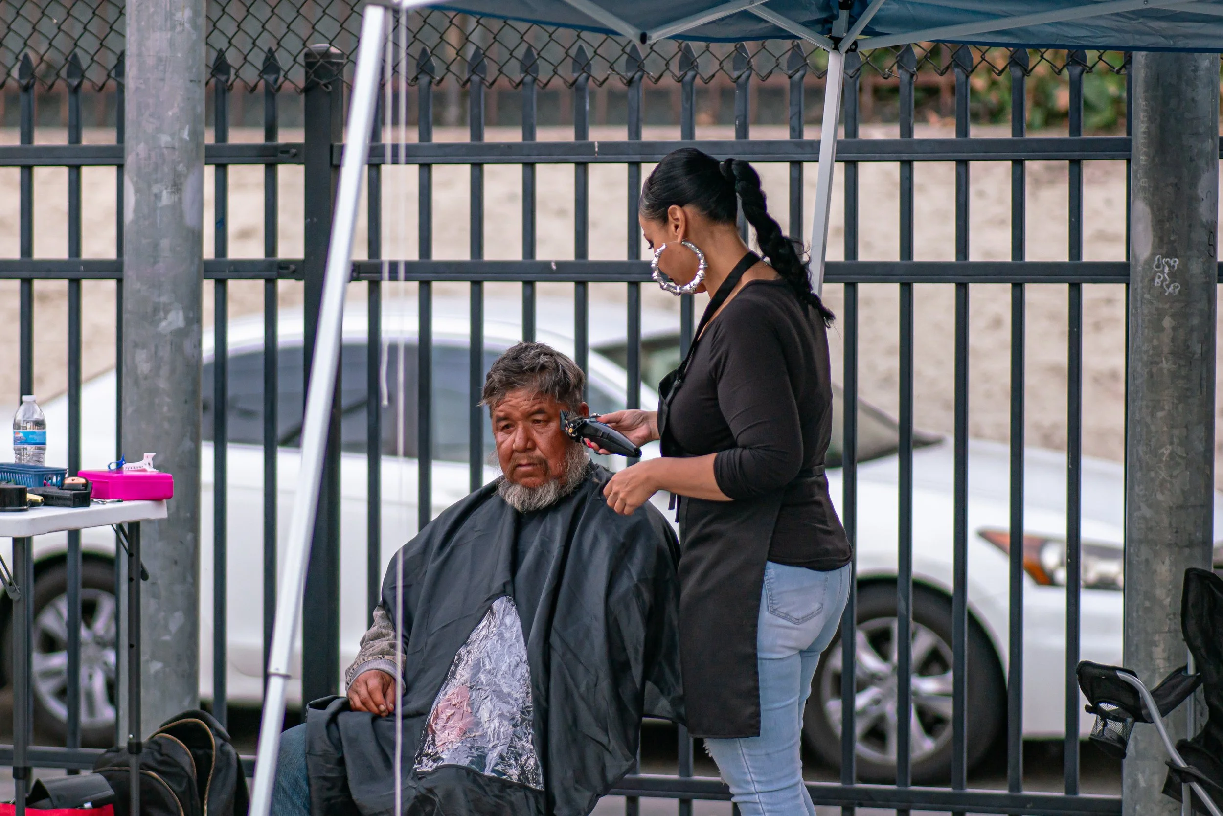 A woman giving a haircut outdoors to a man sitting on a chair, with a black cape around him, in front of a metal fence, with cars parked behind.
