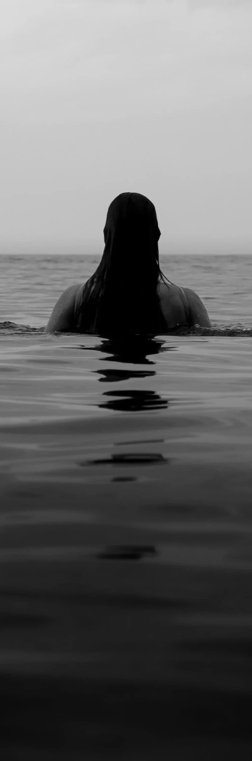 A person with long hair in the water, facing away, with a calm ocean and cloudy sky in the background, captured in black and white.