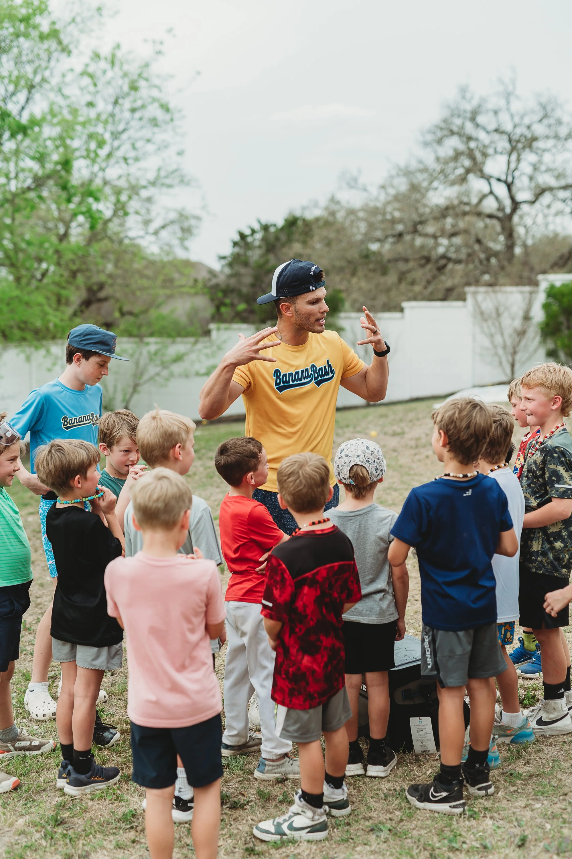 A man wearing a yellow Banana Boat T-shirt and a backward cap is instructing a group of young kids outside in a grassy yard, with trees and a white fence in the background.