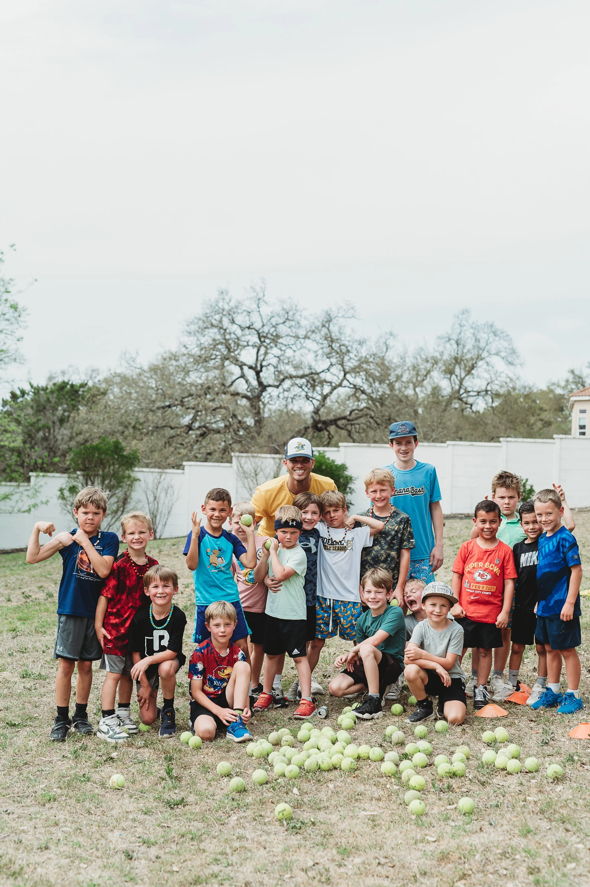 Group of children and two adults standing outdoors on grass field, surrounded by scattered tennis balls, with trees and a white fence in the background.