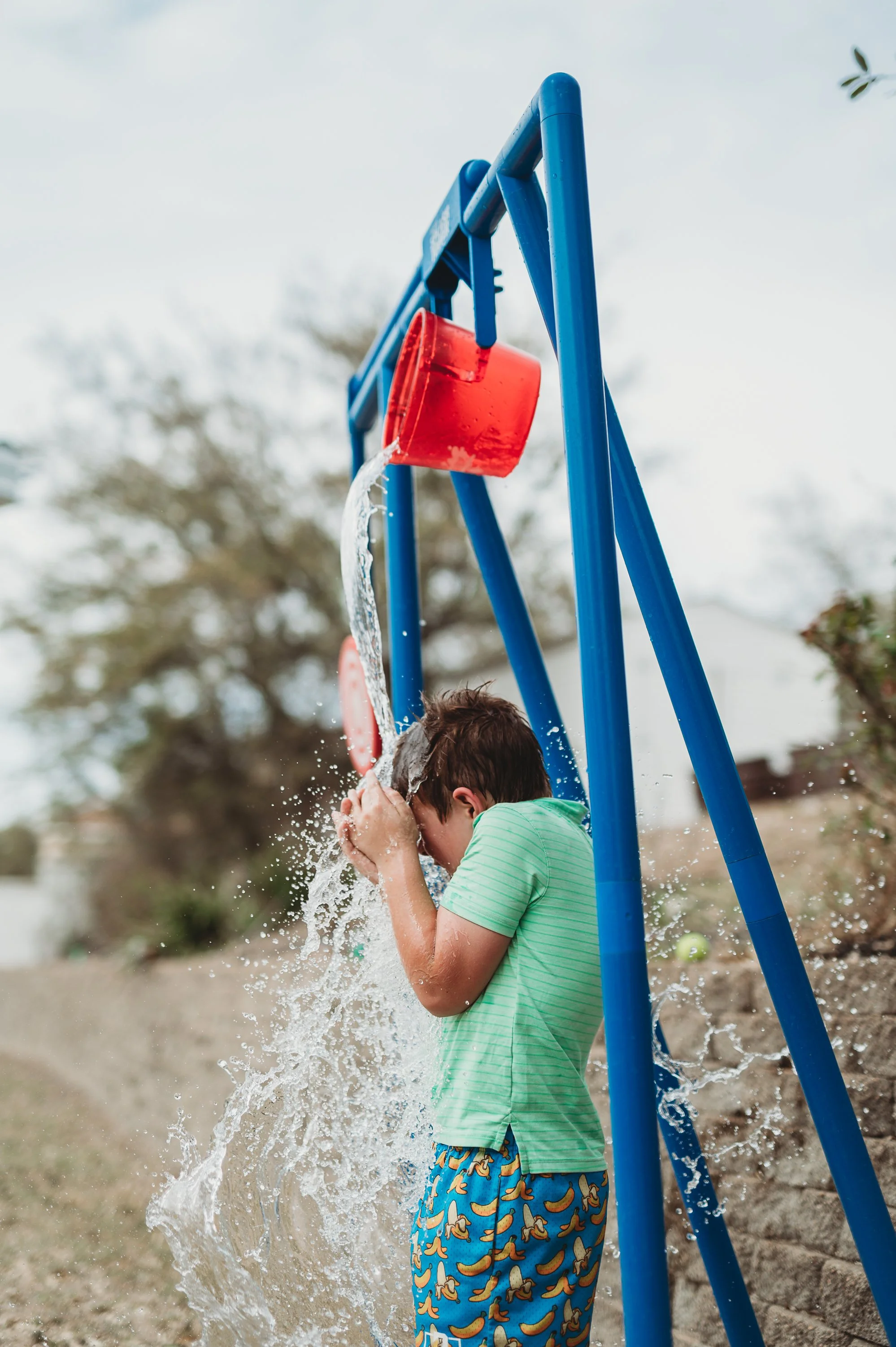 Child playing under a blue swing set, with water pouring from a red bucket.