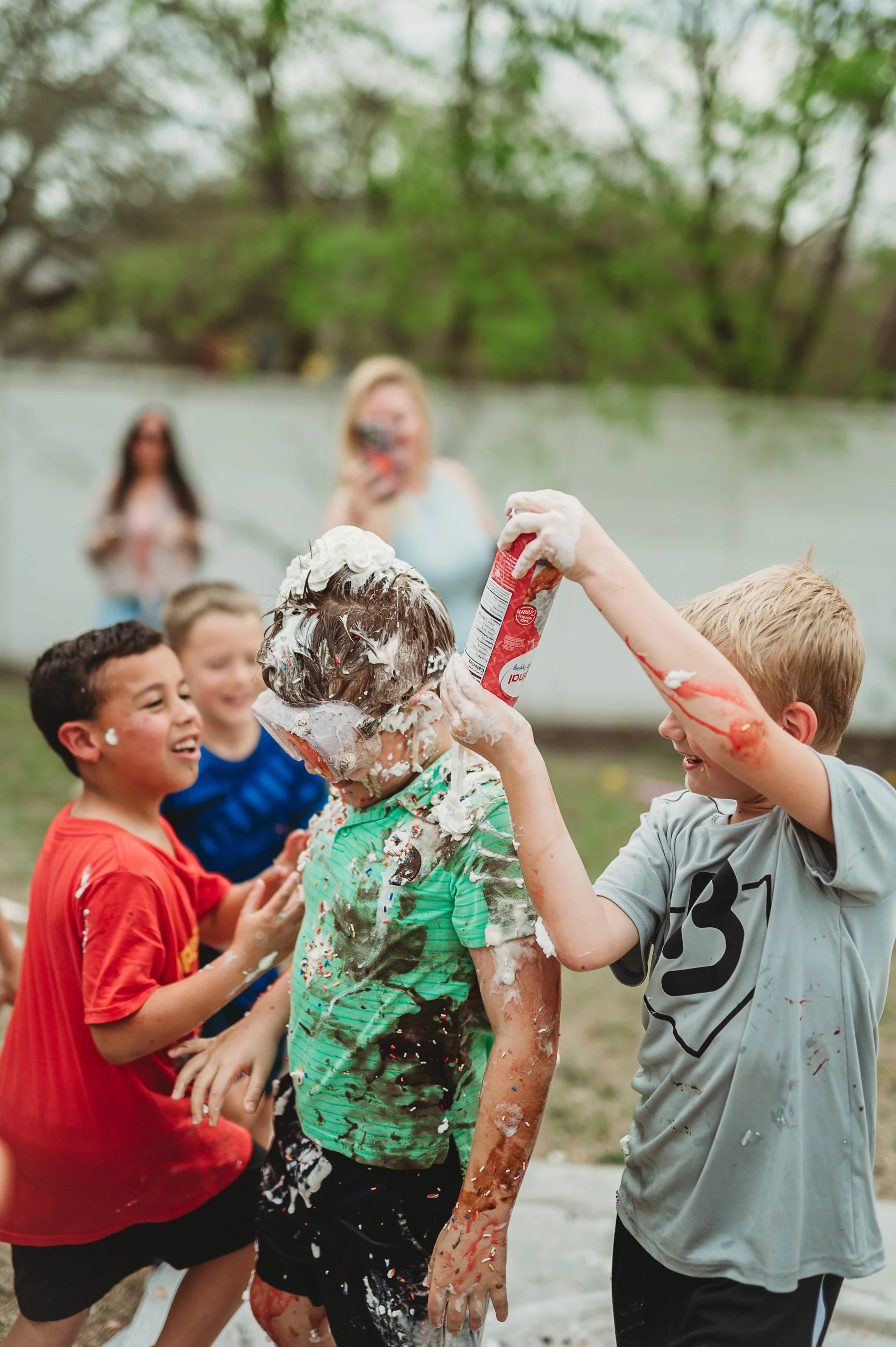 Children at a summer outdoor party, throwing and playing with foam and spray from a can, with adults watching in the background.