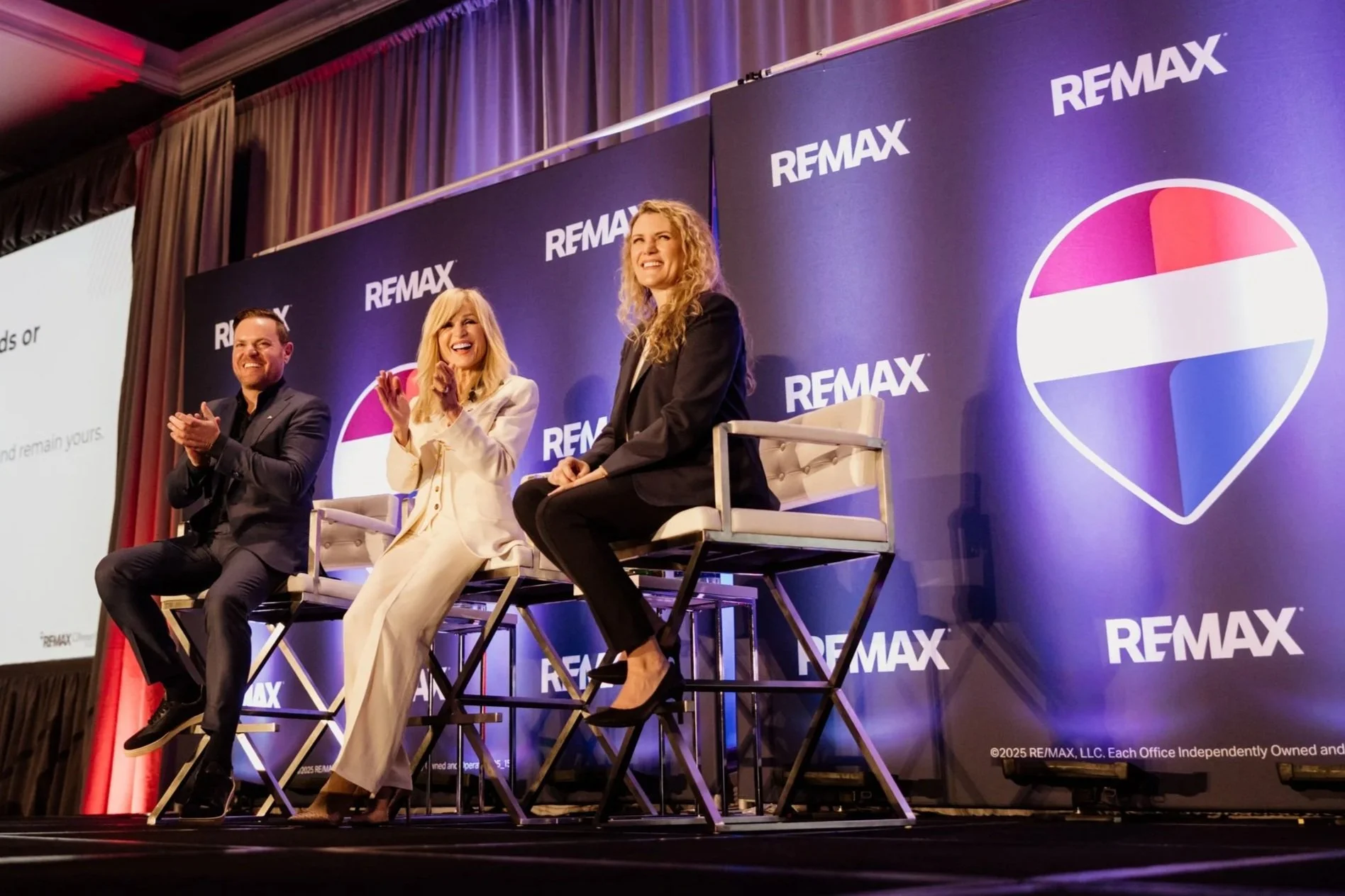 Justin, Vivian & Michelle Risi sit on stage, smiling with a REMAX-branded banner behind them