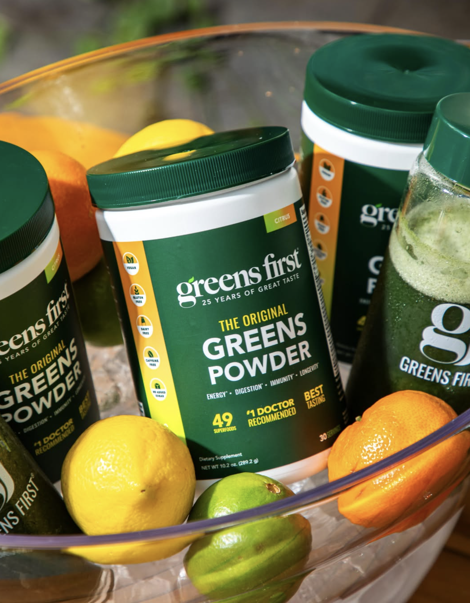 Greens First green powder supplement containers with fresh citrus fruits in a glass bowl