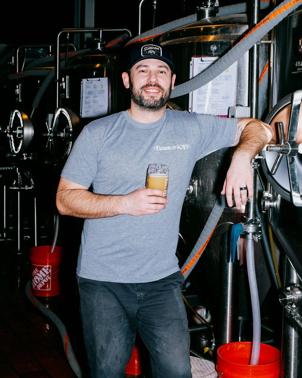 Man with a beard in a gray T-shirt and black cap holding a beer in a brewery or beer brewing facility.